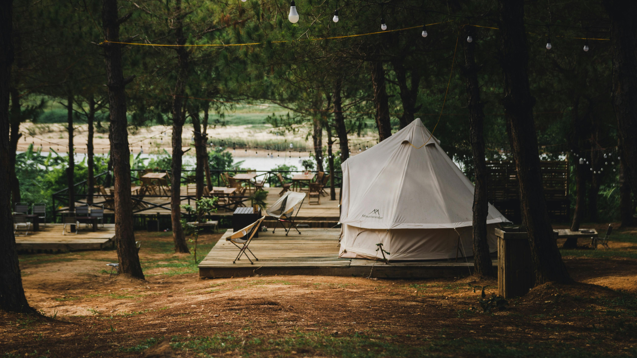 Ett ljust glampingtält på en träbrygga i tallskog vid en lugn sjö, med ljusslingor mellan träden